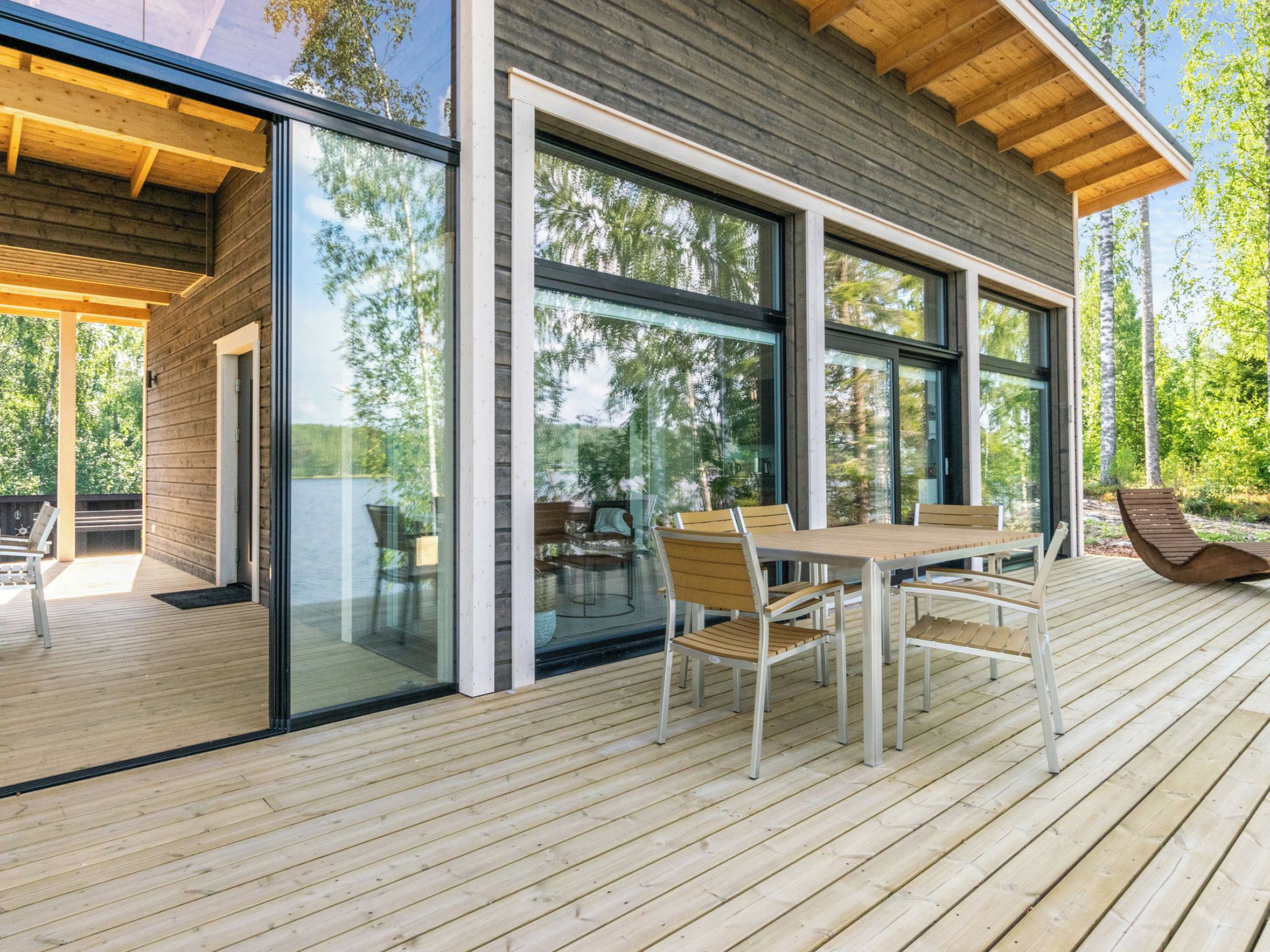 Een modern terras buiten, met een houten tafel en stoelen, grote glasramen en uitzicht op het omliggende bos.