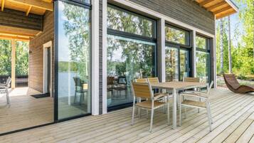 Een modern terras buiten, met een houten tafel en stoelen, grote glasramen en uitzicht op het omliggende bos.