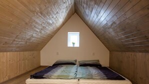 Ein Zimmer mit schrägem Dach, Holzwänden, einem kleinen Fenster und einem Bett mit bunter Decke.