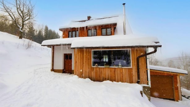 Eine Holzhütte mit schneebedecktem Dach und Schornstein, eingebettet in eine verschneite Winterlandschaft.