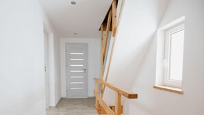 Ein schmaler Flur mit einer Holztreppe auf der rechten Seite, einer weißen Tür am Ende und einem Fenster an der gegenüberliegenden Wand.