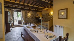 Comedor con una larga mesa dispuesta para cuatro personas, paredes de piedra, vigas de madera y chimenea de piedra.