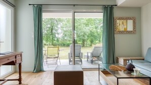 En moderne stue med trægulv, et sofabord i glas, en sofa og et skrivebord. Der er en skydedør af glas, som fører ud til en udendørs terrasse med stole og et bord.