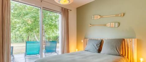 Une chambre dotée d’un grand lit, d’une tête de lit en bois et donnant sur un espace extérieur aménagé avec des chaises, accessible par une porte coulissante.