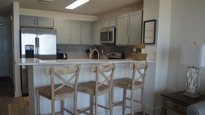 A kitchen with a stainless steel refrigerator, a microwave, and a coffee maker on the countertop. There are three wooden chairs with a unique design. - The Calypso Resort in PCB , Family Friendly,11 Floor, Free Beach Chairs Service (Panama City Beach)