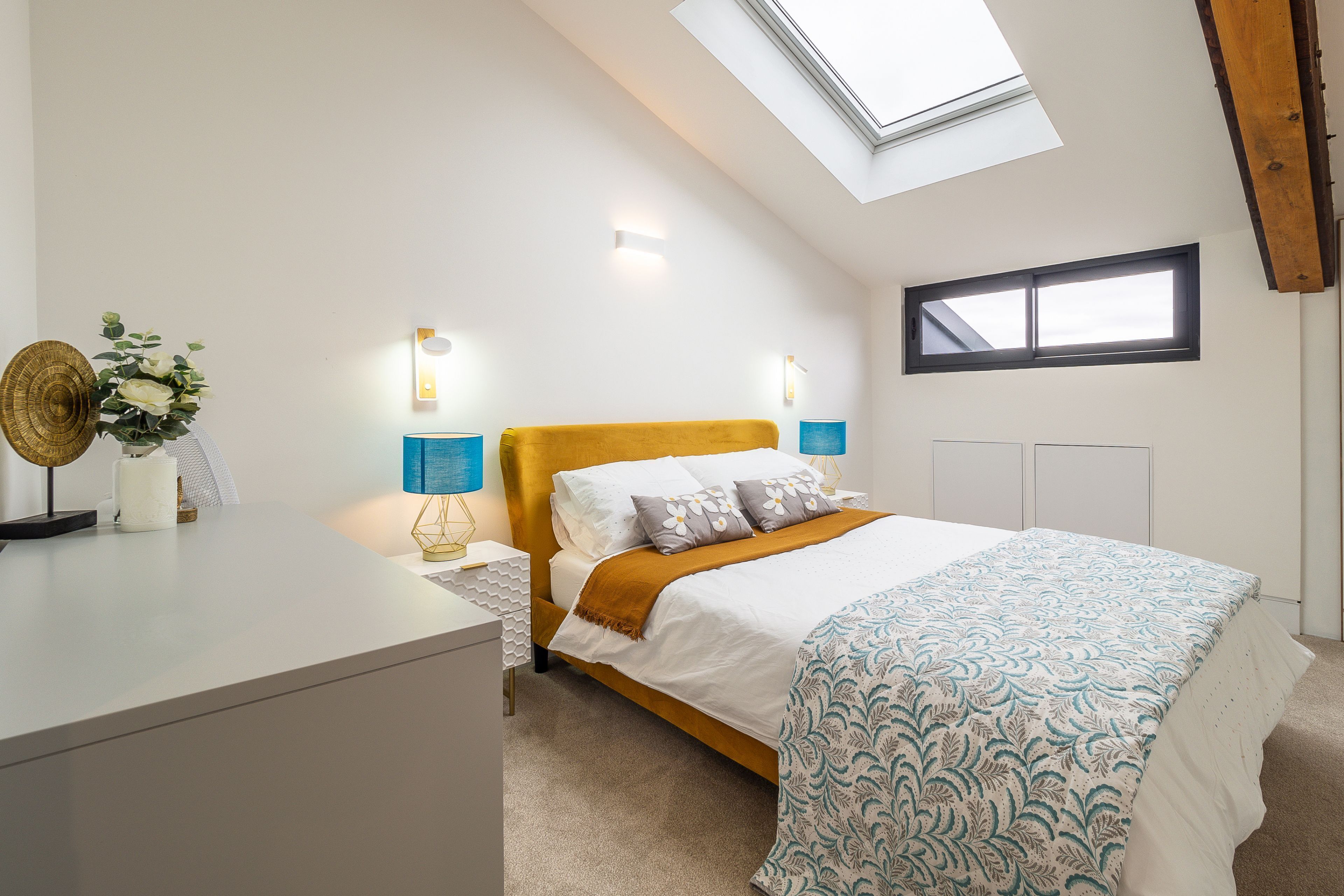 A bedroom with a sloped ceiling, a bed with a white and blue patterned bedspread, two bedside lamps, and a skylight.