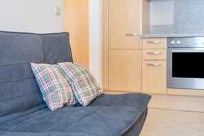 A grey sofa with plaid cushions, a kitchen with wooden cabinets and a stainless steel oven, and a tiled floor.
