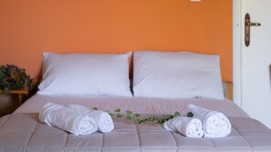 Un lit avec du linge de lit blanc et deux serviettes enroulées, une plante en pot et un mur orange.