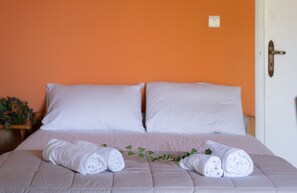 Un lit avec du linge de lit blanc et deux serviettes enroulées, une plante en pot et un mur orange.