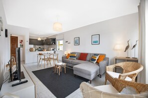 A modern living room with a grey sofa, wicker chairs, a dining area, and a kitchen in the background.