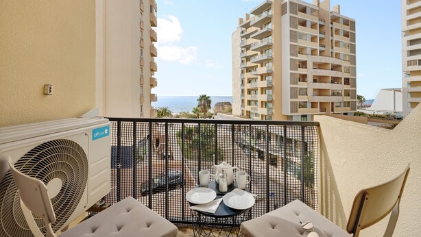 A balcony with a table set for two, a fan, and a view of the sea and other buildings. - Apartment "Casa Farol" with Sea View, Balcony & Wi-Fi (Portimão)