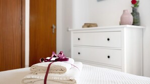 A neatly made bed with folded towels and a ribbon, a white dresser, and a wooden door in a room. - Apartment "Casa Farol" with Sea View, Balcony & Wi-Fi (Portimão)
