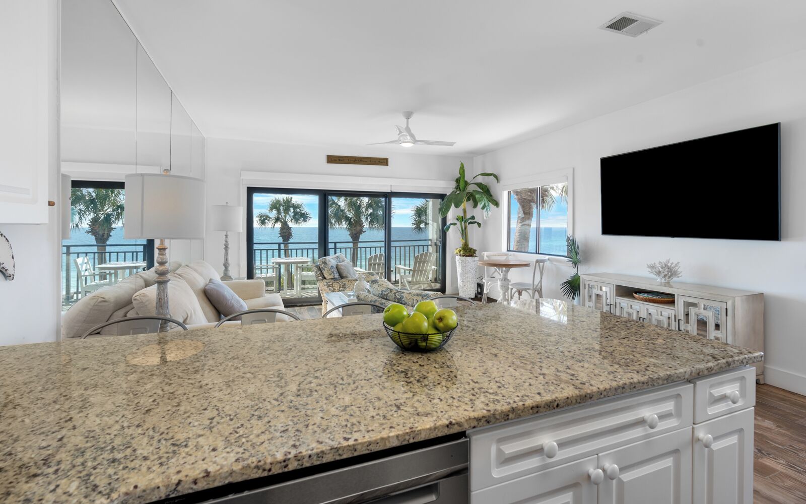 A modern kitchen with granite countertops, a bowl of green apples, and a view of the ocean through large windows.