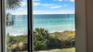 A view of the ocean through a window, with a grassy area and some plants in the foreground. - Beachfront Condo + Bikes, Community Pool & Views (Santa Rosa Beach)
