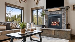 Ein gemütliches Wohnzimmer mit Kamin aus Stein, großen Fenstern, einer Couch mit gemusterten Kissen und einem Couchtisch mit Dekoration.