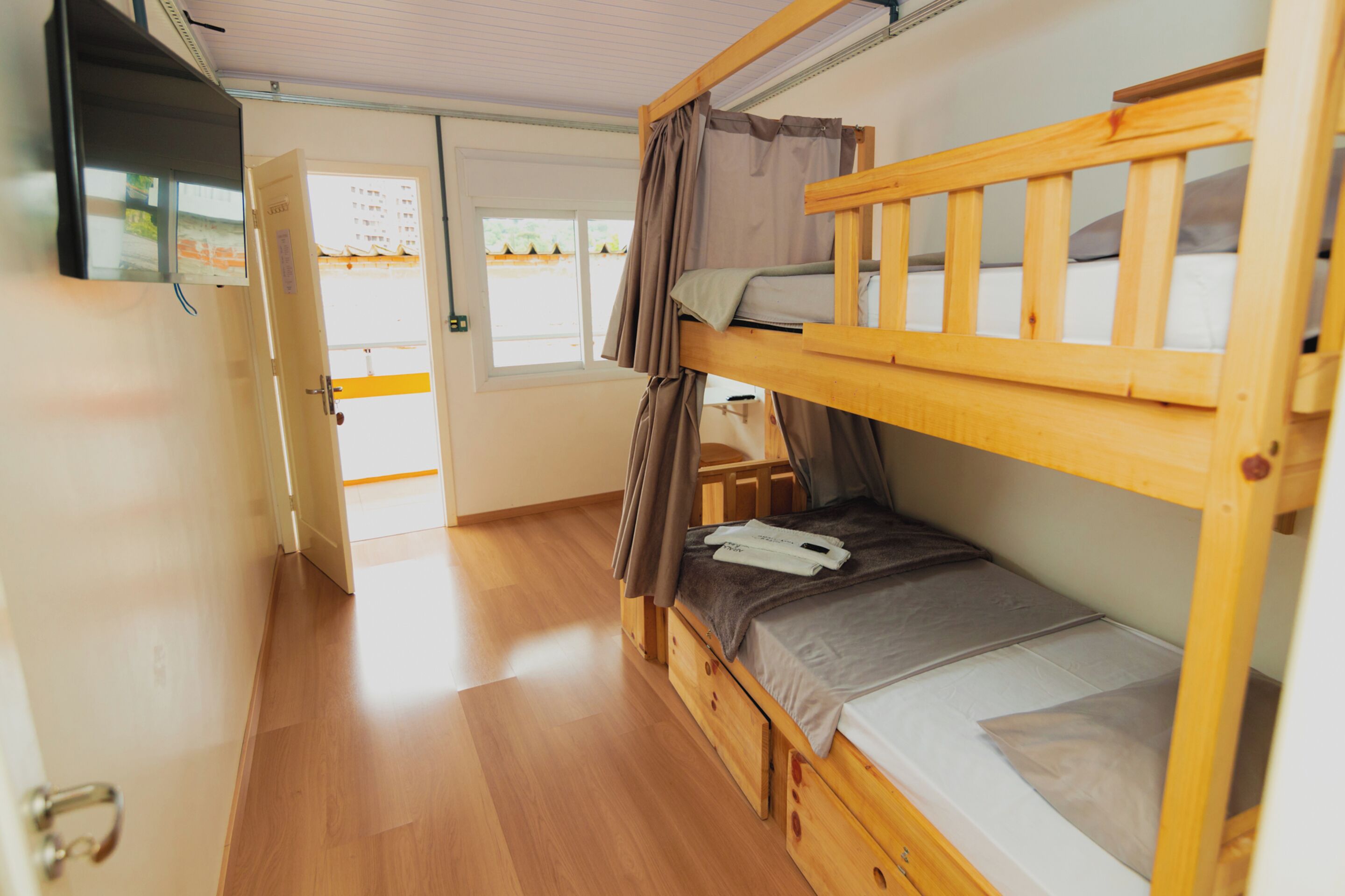 Habitación con literas, suelo de madera, televisión montada en la pared y puerta que da a un espacio exterior.