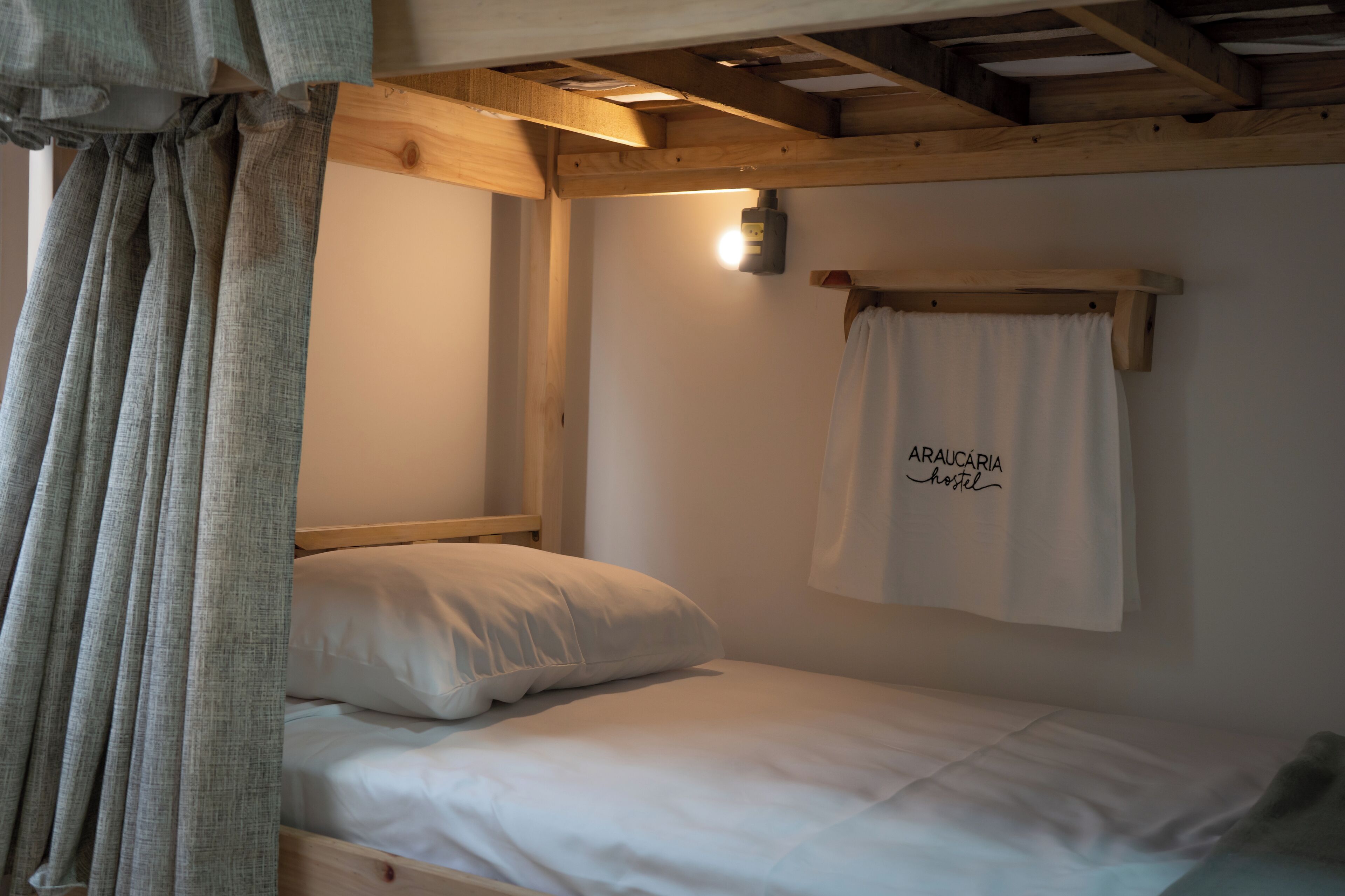 A single bed with a wooden headboard, a white pillow, and a white curtain with "Arauçaria Hostel" written on it.