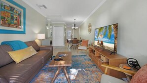A living room with a brown leather sofa, a coffee table, a TV on the wall, and a dining area in the background.