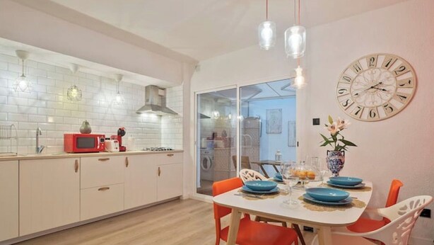 Una cocina moderna con armarios blancos, un microondas rojo y un comedor con mesa blanca y sillas naranjas.