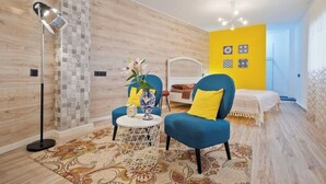 Un dormitorio moderno con piso de madera, una cama con cabecera blanca, un sillón azul, una pequeña mesa redonda con un florero y una lámpara de pie.