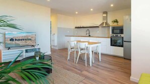 Una cocina moderna con armarios blancos, una encimera de madera y electrodomésticos de acero inoxidable. Hay un comedor con sillas blancas y una mesa pequeña. Un televisor está montado en la pared, y se ve una planta en primer plano.