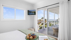 A room with a bed, a dining table with pastries and a cup, and a balcony with a view of the sea and mountains.