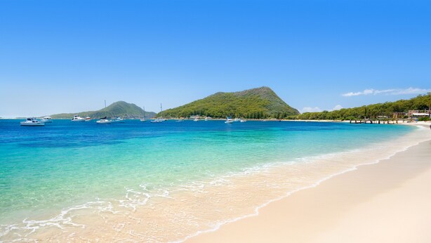 A beach with clear turquoise water, boats, and a mountain in the background.