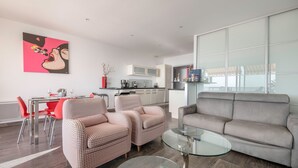Ein modernes Wohnzimmer mit einem grauen Sofa, zwei gemusterten Sesseln, einem Glastisch und einem Essbereich mit roten Stühlen und einem Glastisch.