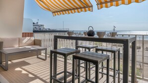 Ein Balkon mit einer Couch, einem Tisch und Stühlen, mit Blick auf ein Gewässer und eine entfernte Küstenlinie.