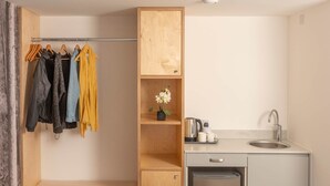 Een klein keukenhoekje met een spoelbak, een kleine koelkast en een koffiezetapparaat. Er is een houten plank met een potplant en een kledingrek met verschillende kledingstukken.