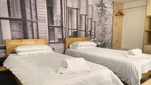 Deux lits simples avec du linge de lit et des serviettes blancs, des têtes de lit en bois et une grande fenêtre donnant sur des arbres et des bâtiments.