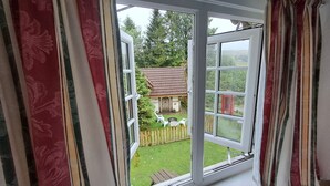Une vue à travers une fenêtre avec des rideaux, dévoilant une maison avec un toit en tuiles, une clôture en bois et une cour verdoyante.