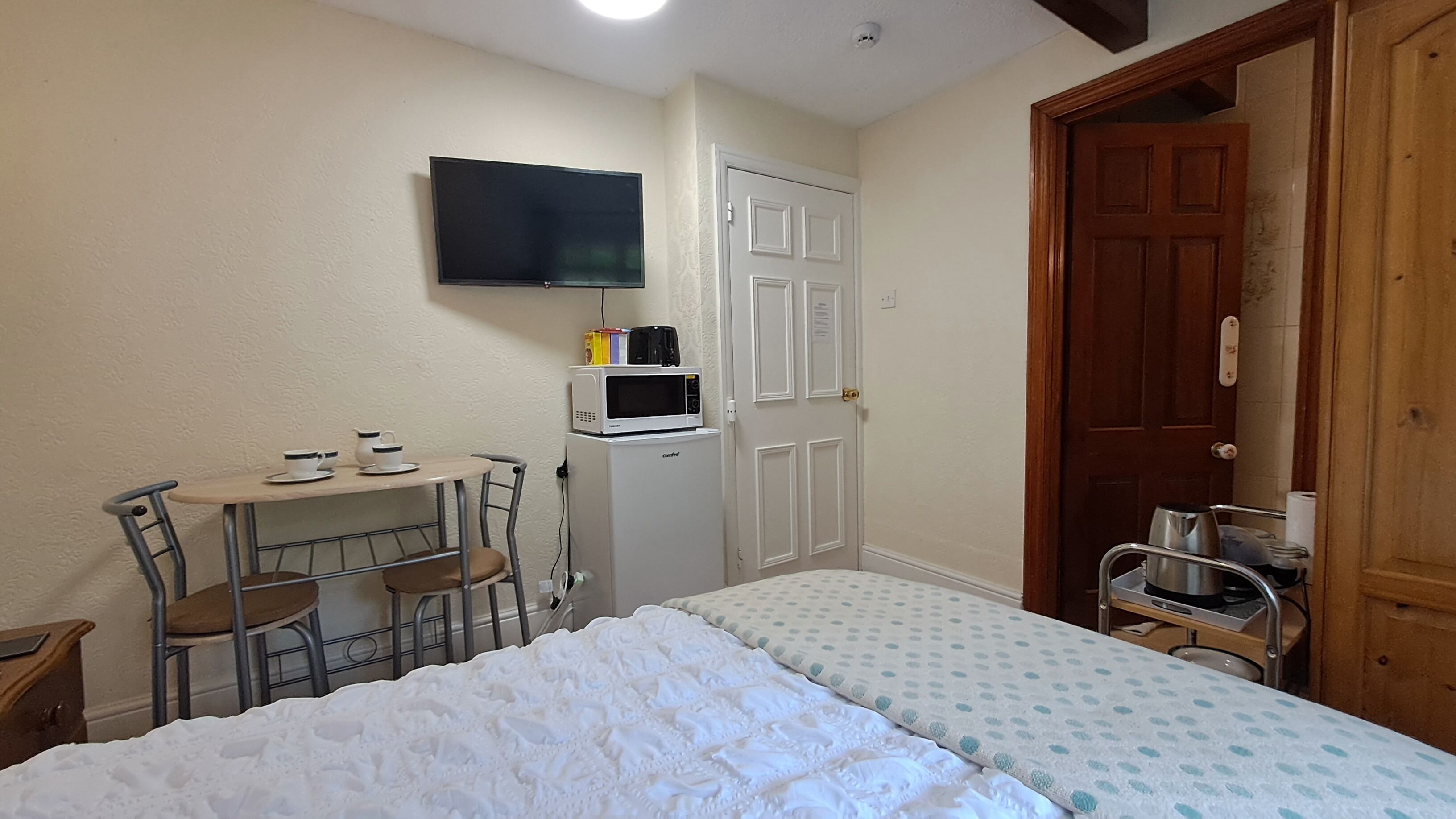 Una habitación pequeña con una cama, un escritorio con una silla, un microondas y un televisor en la pared.