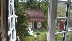 On aperçoit, à travers une fenêtre, un petit jardin clos, avec un bâtiment au toit rouge, des chaises blanches et une cabine téléphonique rouge.