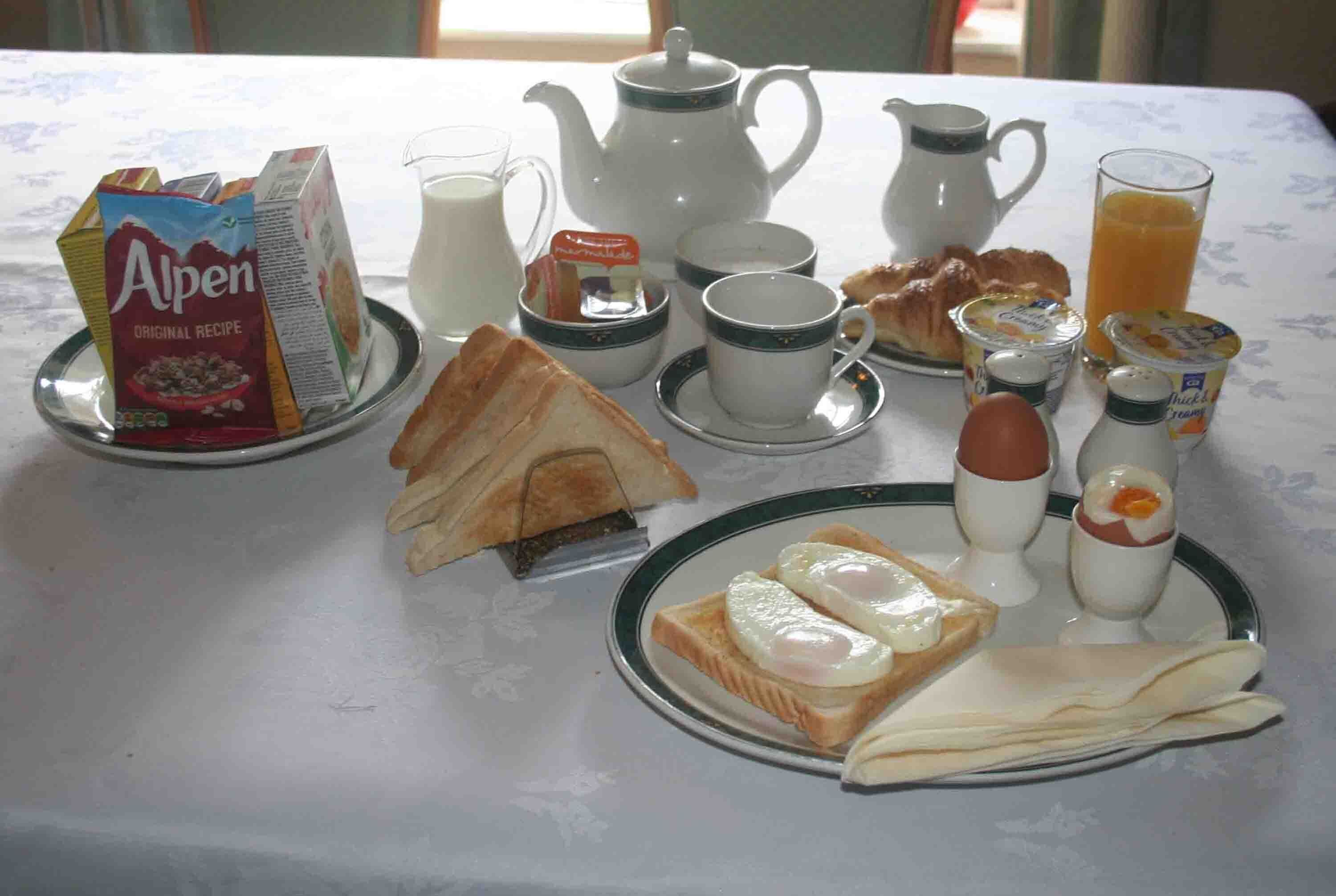 Mesa de café da manhã com leite Alpen em caixa, um bule, xícaras, jarra de leite, torradas, ovos, manteiga e um copo de suco de laranja.