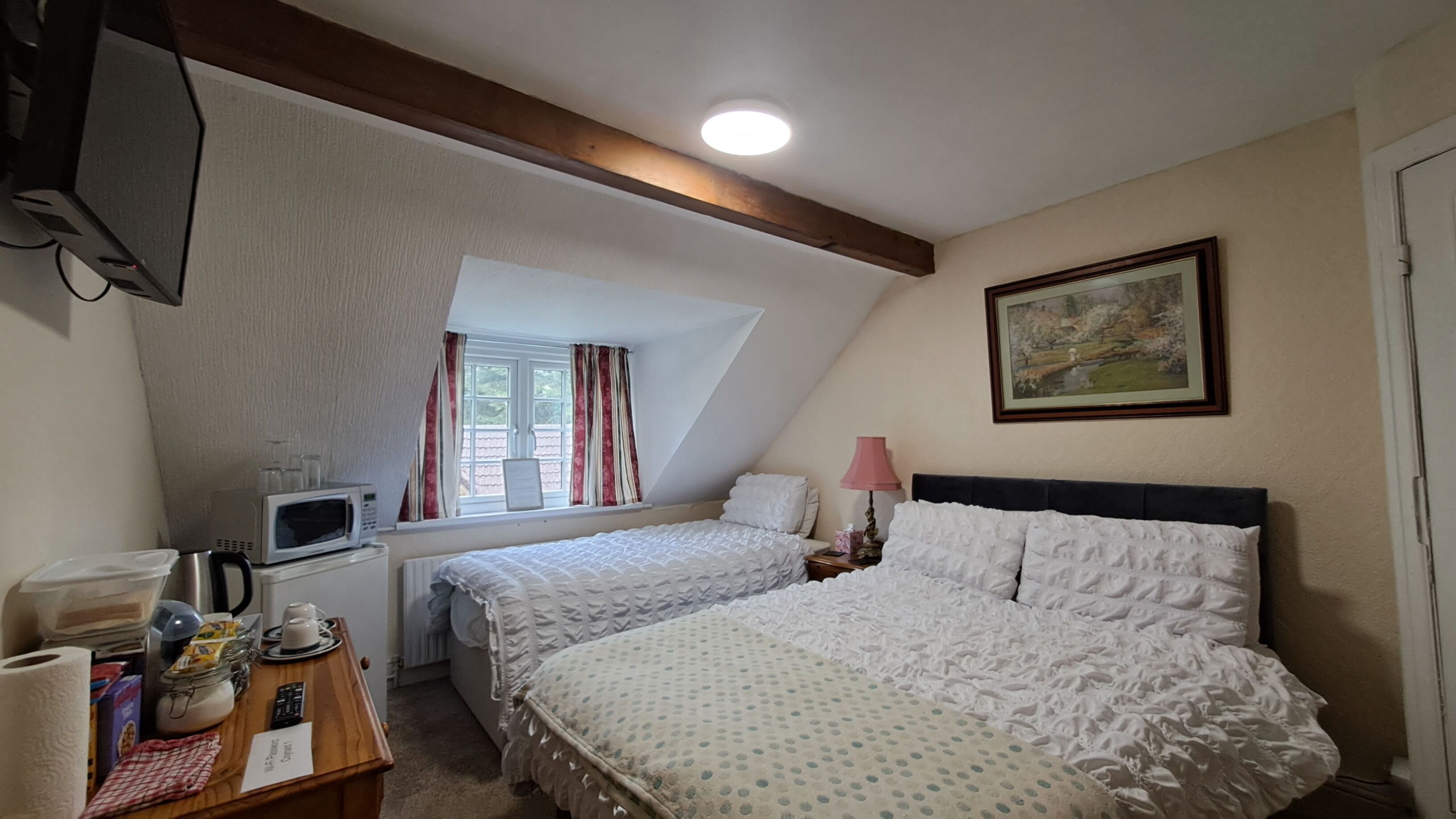 Quarto com duas camas, janela com cortinas, um quadro na parede e uma escrivaninha com micro-ondas e outros objetos.
