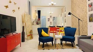 Una sala de estar con televisor, aparador rojo, sofá azul, almohada amarilla y una zona de comedor con mesa y sillas rojas.