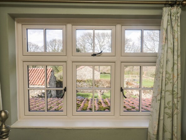 A window with six panes, a curtain, and a view of a garden and buildings. - Bute Cottage (Pickering)
