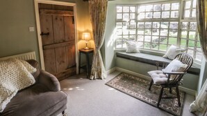A cozy room with a wooden door, a lamp on a small table, a cushioned chair by a window with a view of greenery, and a radiator under a sofa. - Bute Cottage (Pickering)