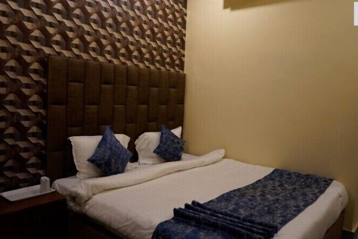 A hotel room with a patterned wall, a tufted headboard, a bed with white linens and blue pillows, and a blue throw blanket.