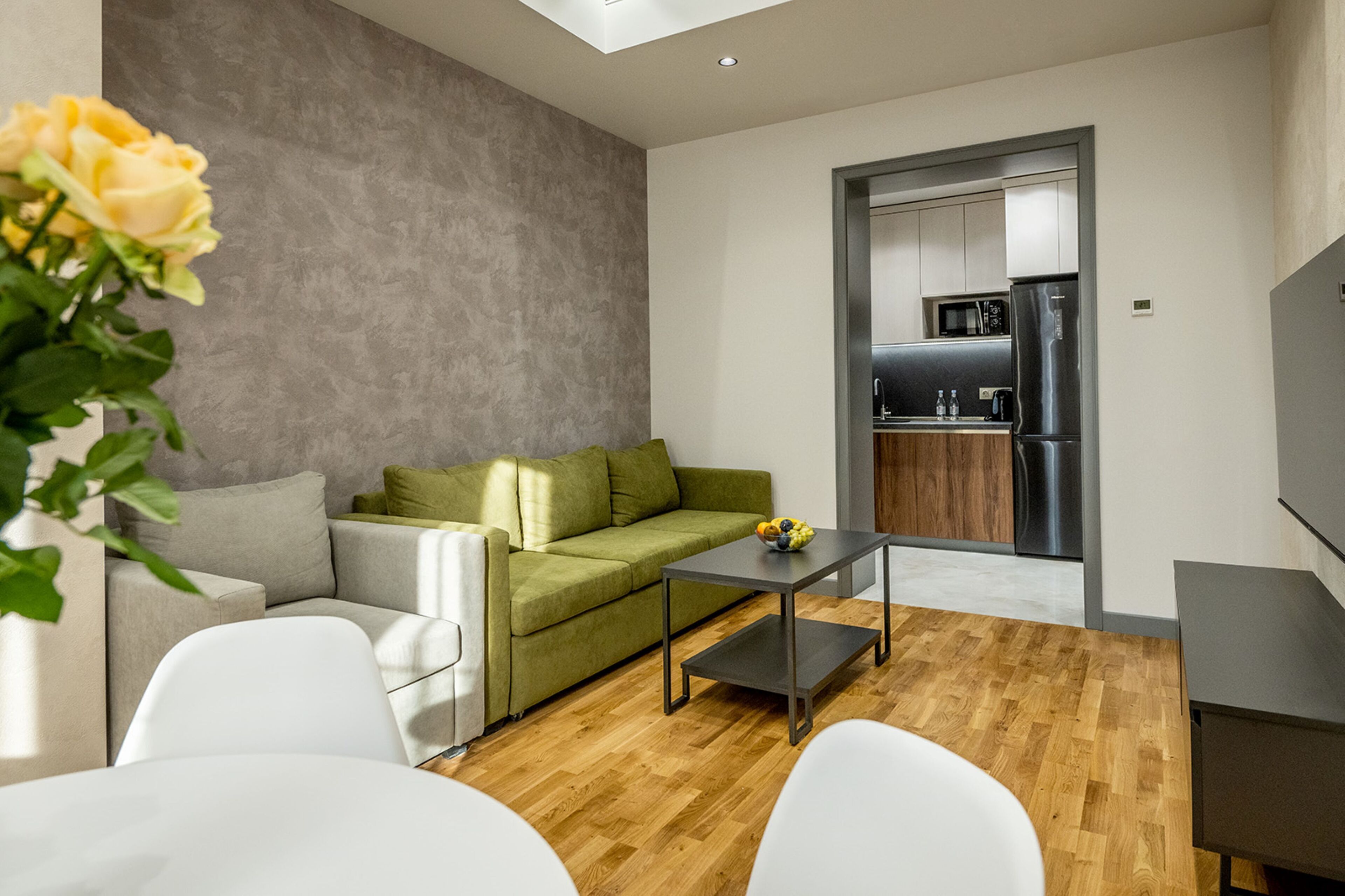 Ein modernes Wohnzimmer mit einem grünen Sofa, weißen Stühlen, einem Couchtisch und einem Fernseher. Durch einen offenen Durchgang ist eine Küche sichtbar.