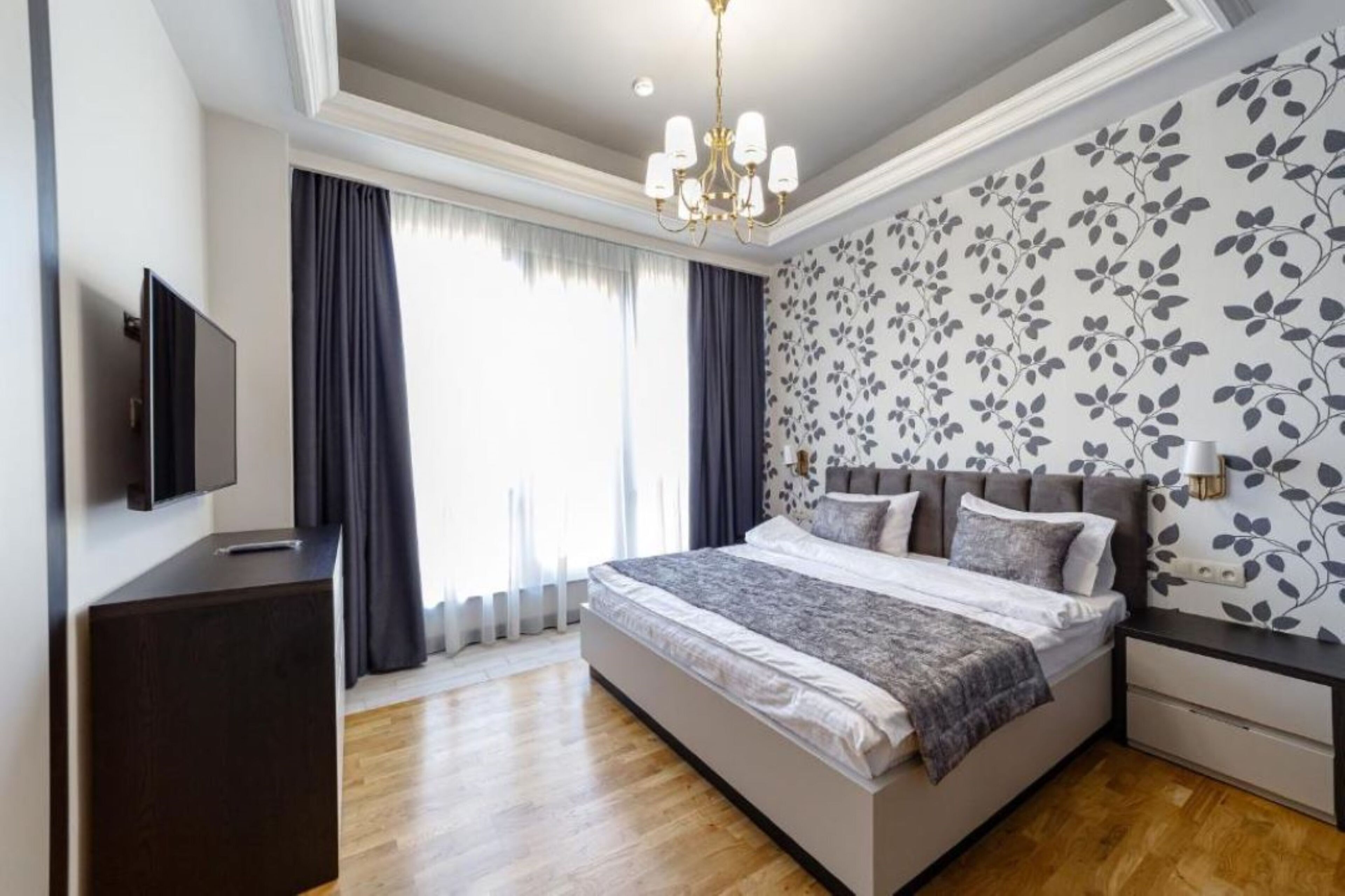 A modern bedroom with a large bed, a flat-screen TV, a dark wooden dresser, and a floral-patterned wall.