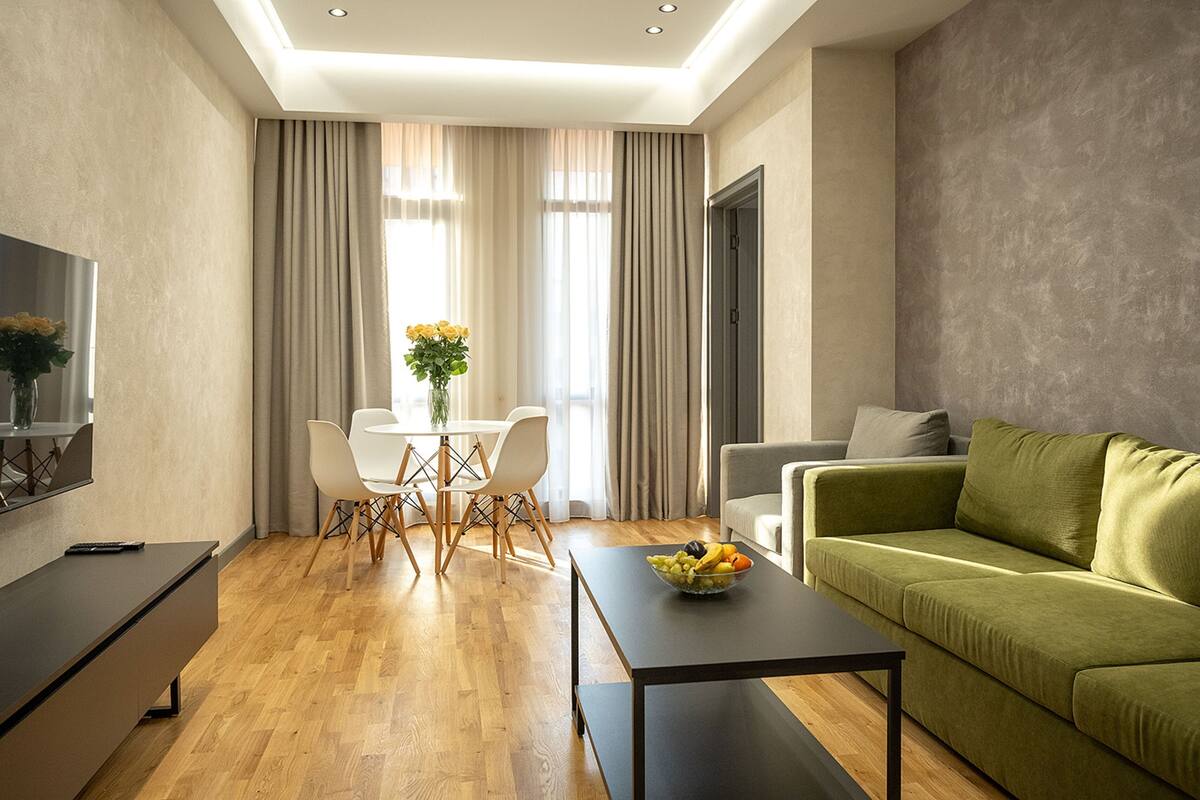 Ein modernes Wohnzimmer mit einem grünen Sofa, einem Esstisch mit Stühlen, einem schwarzen Couchtisch mit Obst und einem Fernseher links.