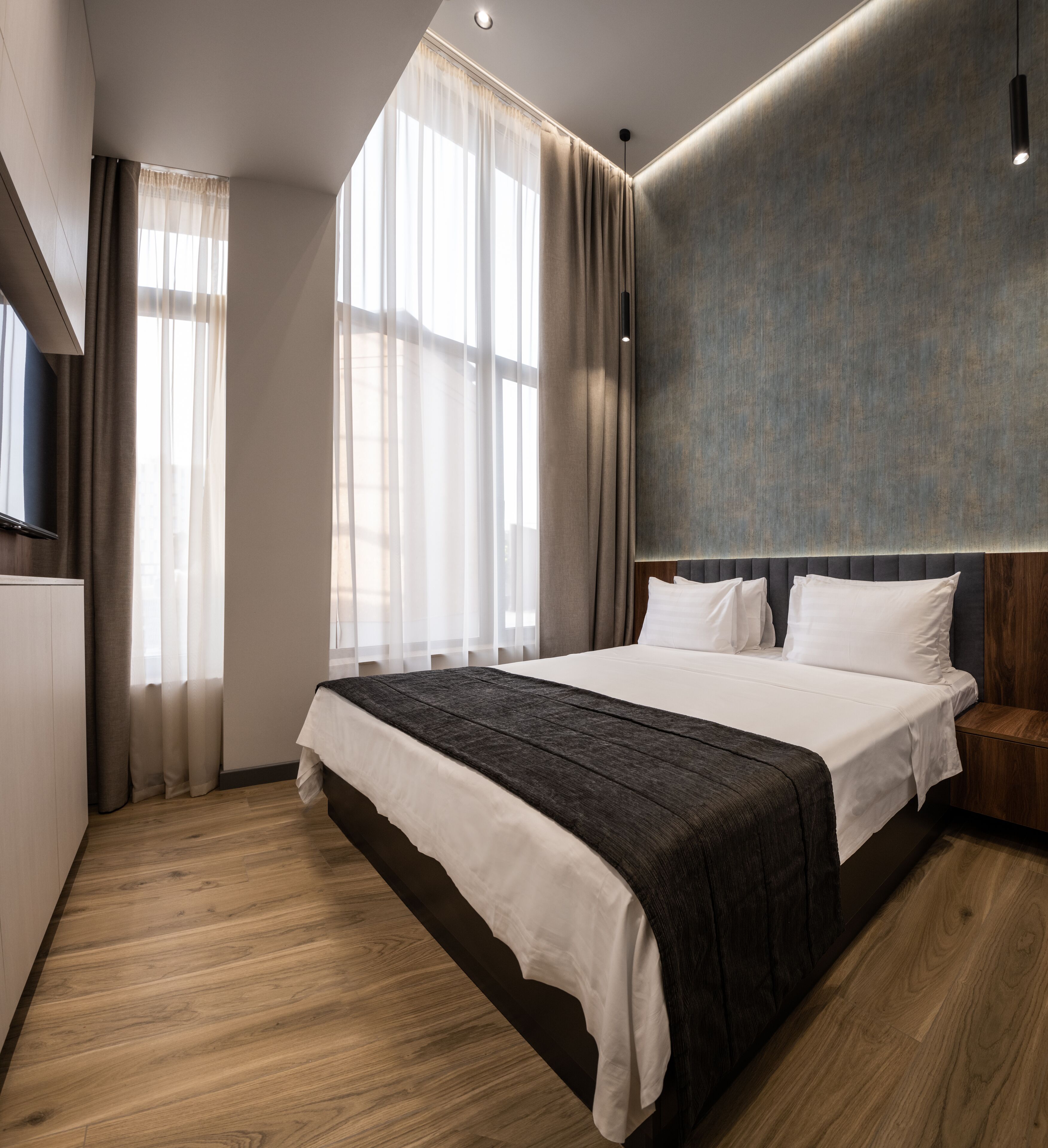 A modern hotel room with a large bed, wooden flooring, and a large window with curtains.