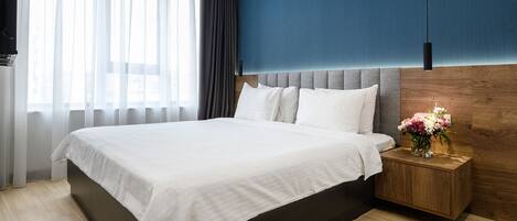 A modern hotel room with a large bed, a wooden headboard, a nightstand with a vase of flowers, and a view of the city through sheer curtains.