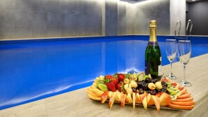 Una bandeja de frutas con una botella de champán y dos copas sobre una mesa en una habitación con piscina azul.