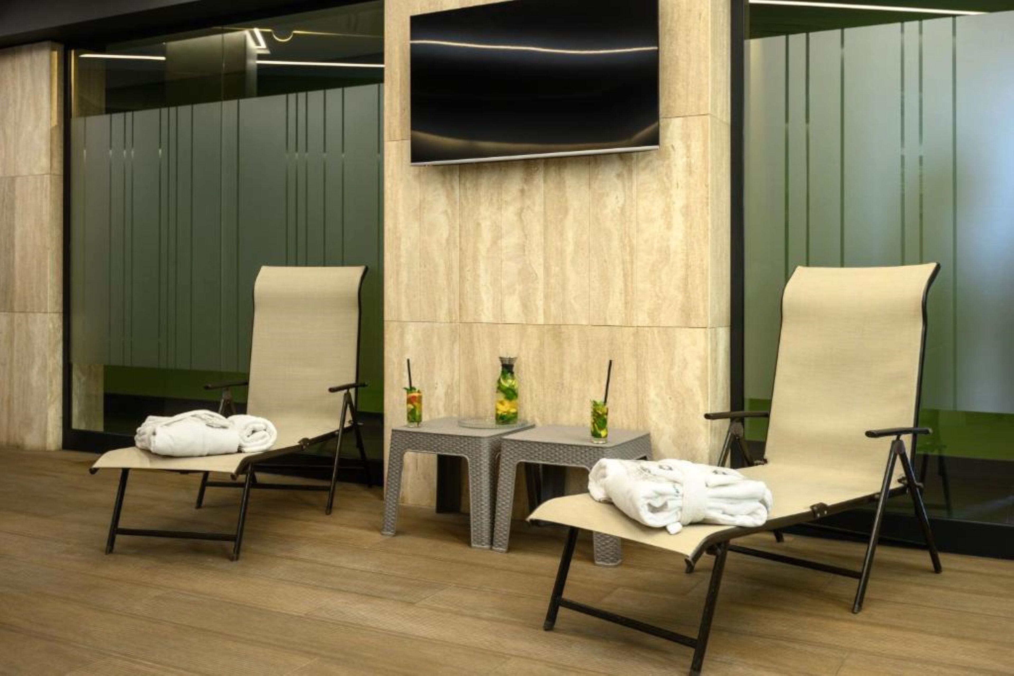 A modern spa room with two reclining chairs, a small table with a vase and a drink, and a flat-screen TV mounted on the wall.