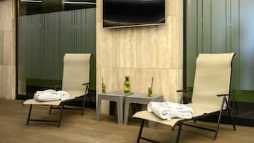 A modern spa room with two reclining chairs, a small table with a vase and a drink, and a flat-screen TV mounted on the wall.