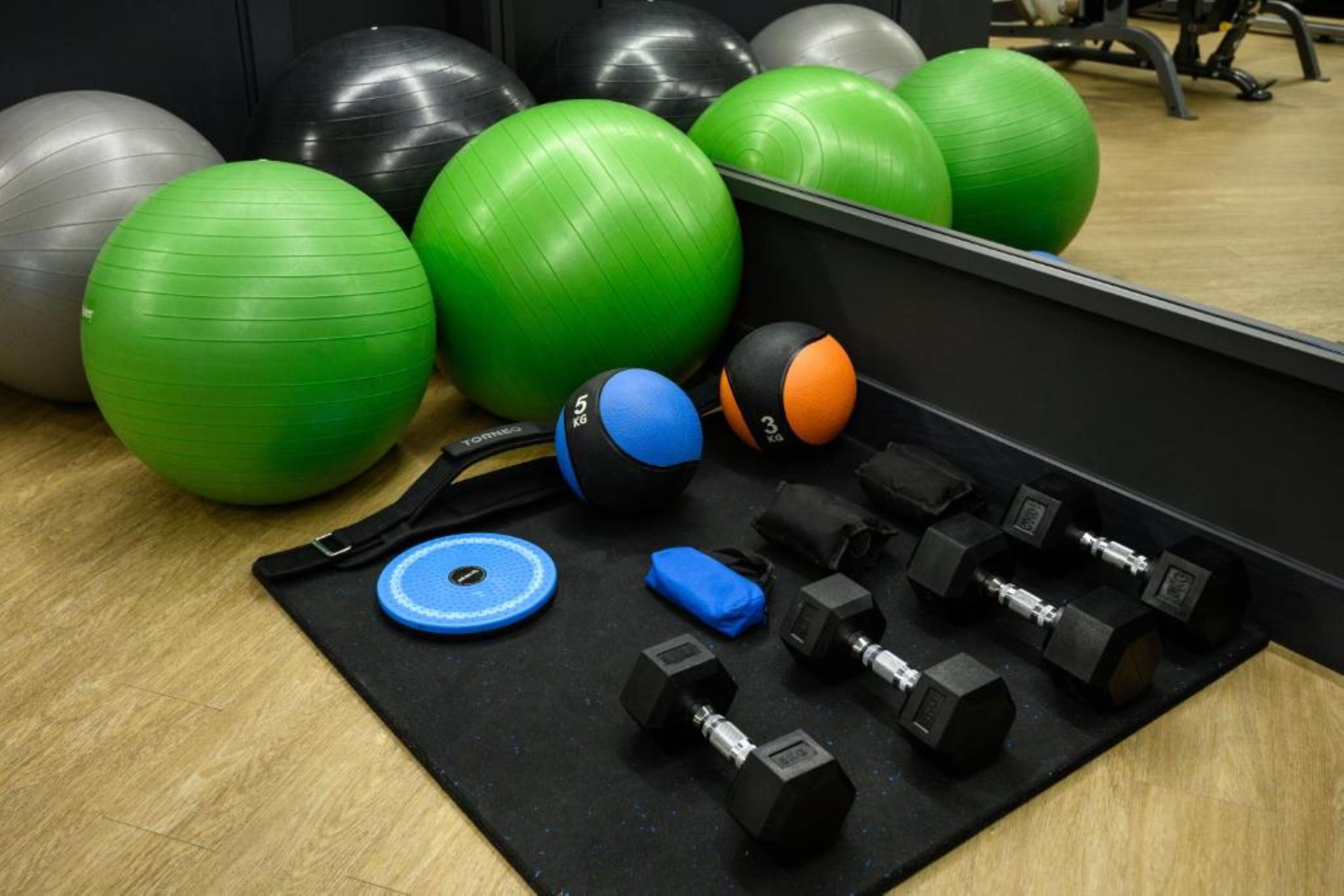 A gym with exercise equipment including large green and black exercise balls, blue and orange medicine balls, and black dumbbells.