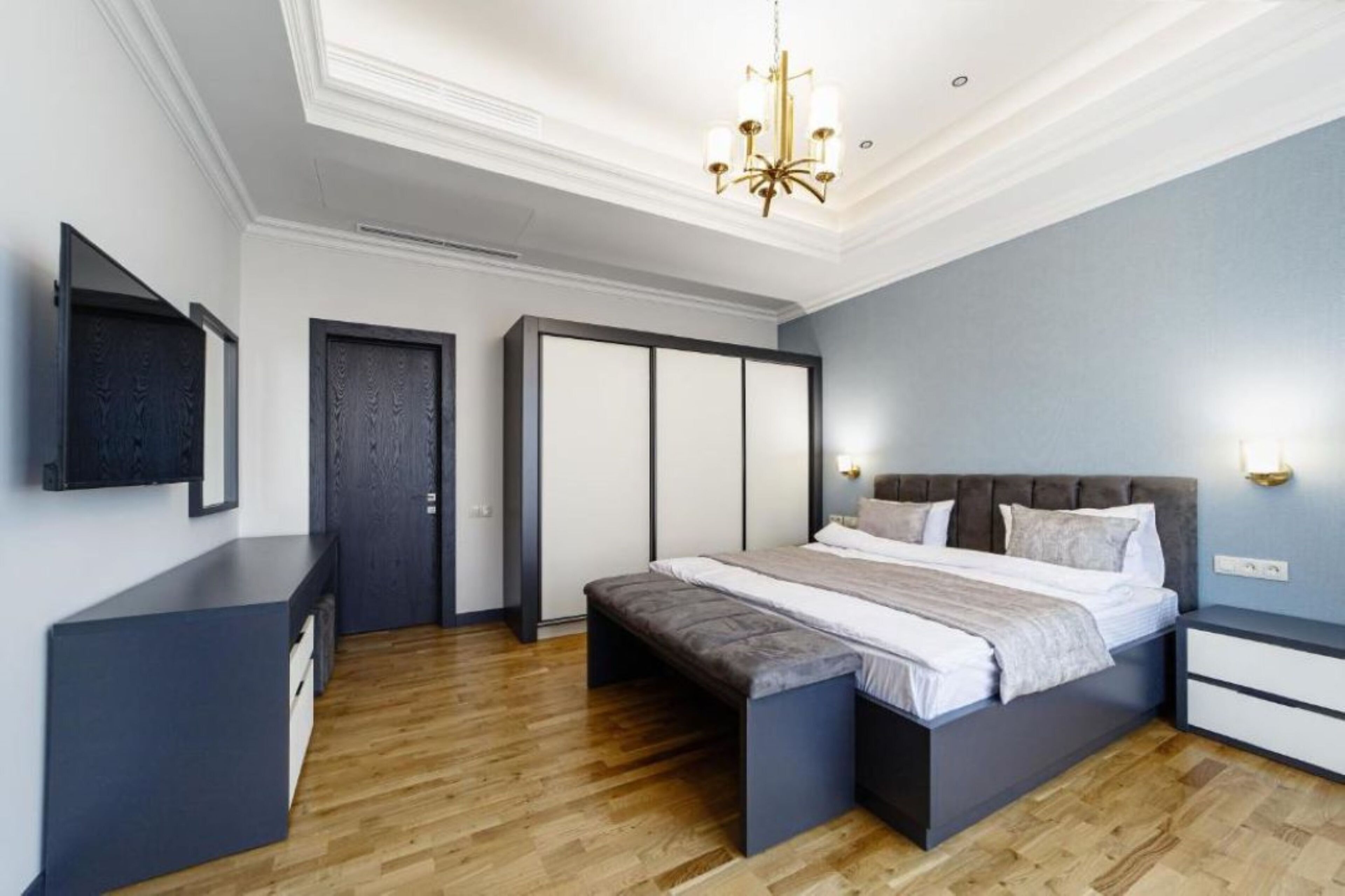 A modern bedroom with a large bed, a bench, a wardrobe, and a TV mounted on the wall.